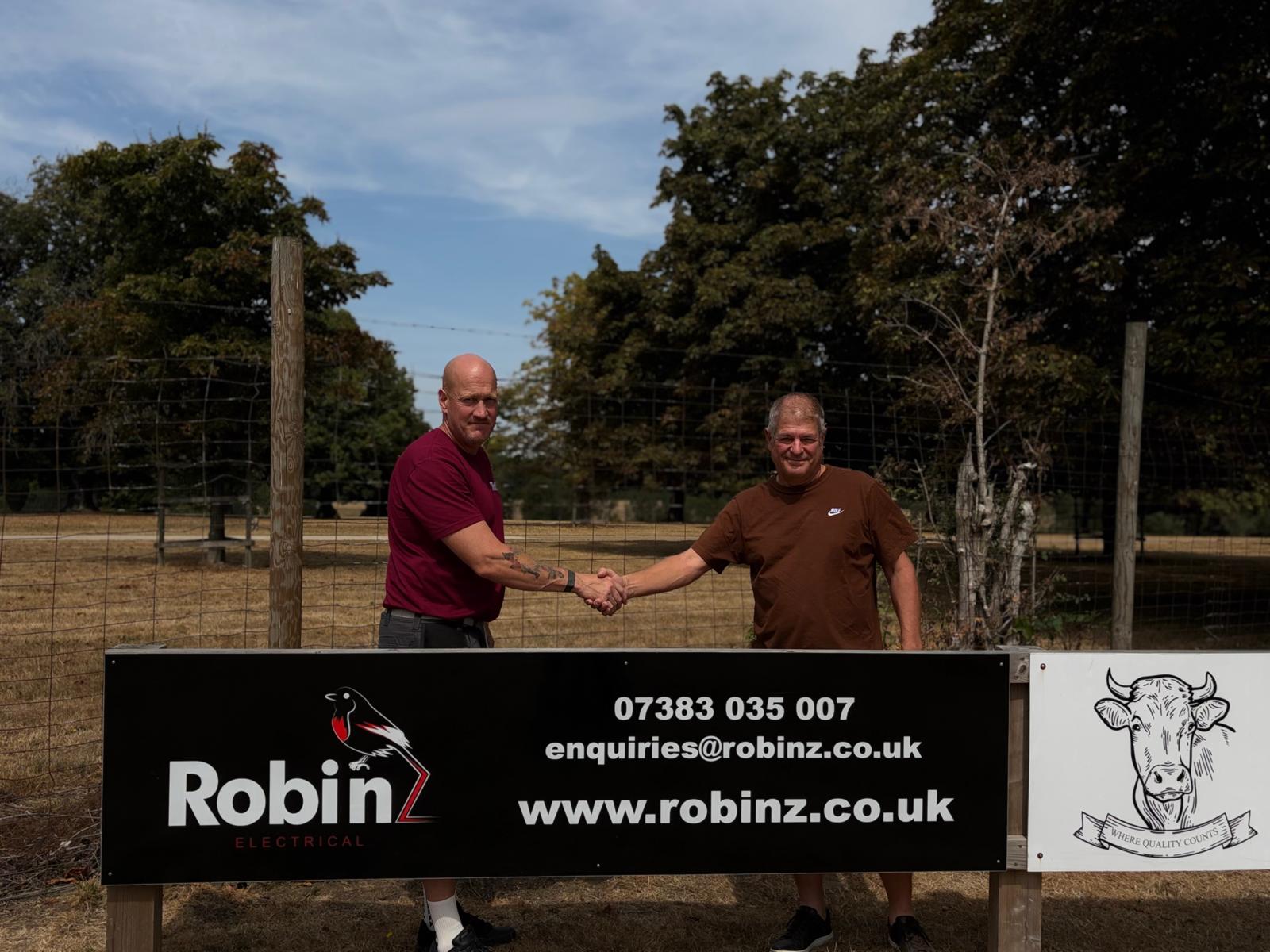 Robinz Electrical founder Alex shaking hands with Knebworth Park Cricket Club chairman Miles Baron by the Robinz Electrical sponsor board